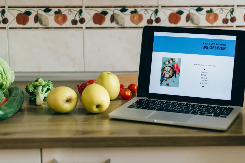 Laptop menampilkan aplikasi pesan antar makanan di meja dapur dengan sayuran segar, gambaran bisnis makanan online